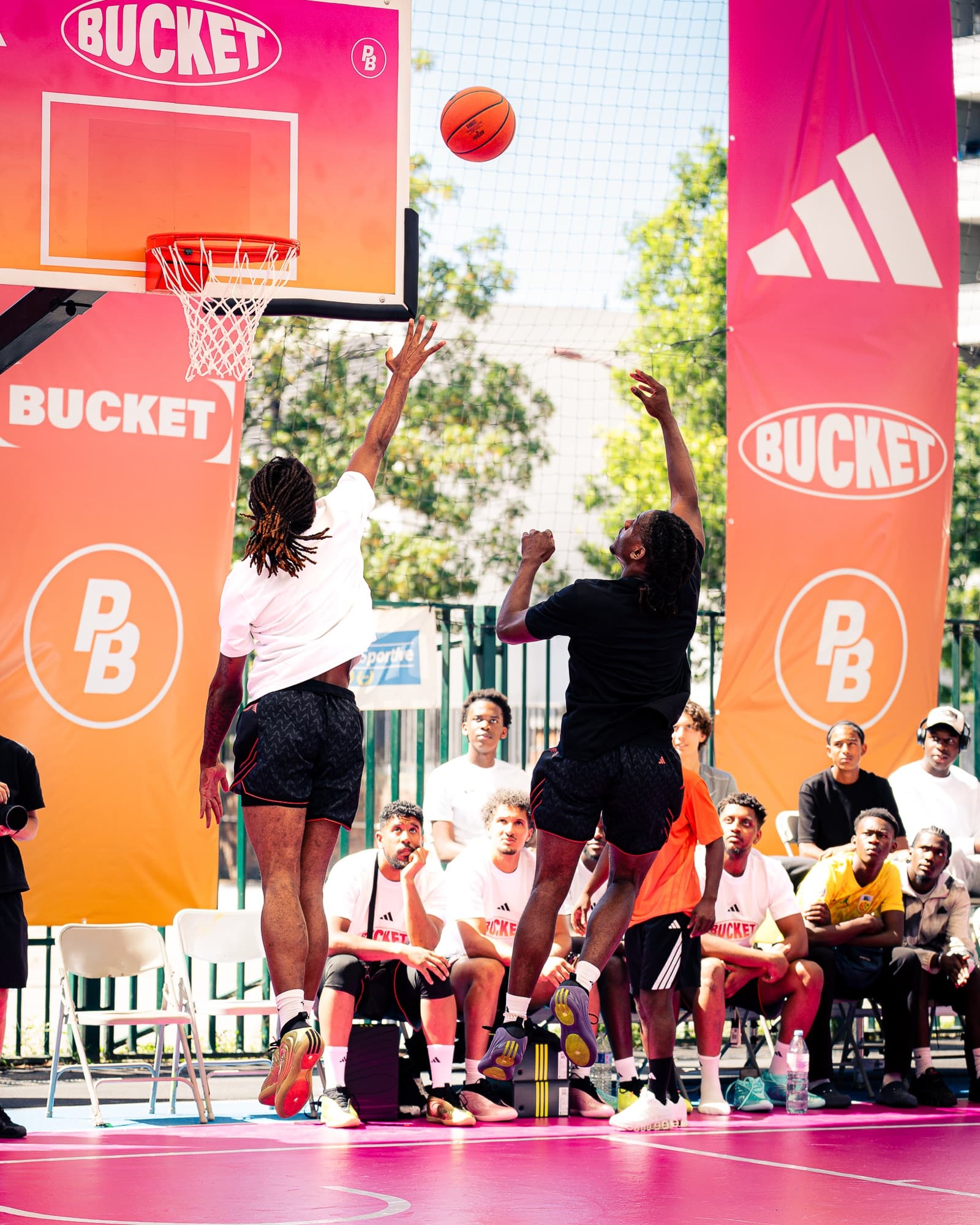 Deux joueurs en suspension près du cercle, devant les bâches orange et roses adidas × BUCKET, public assis au bord du terrain