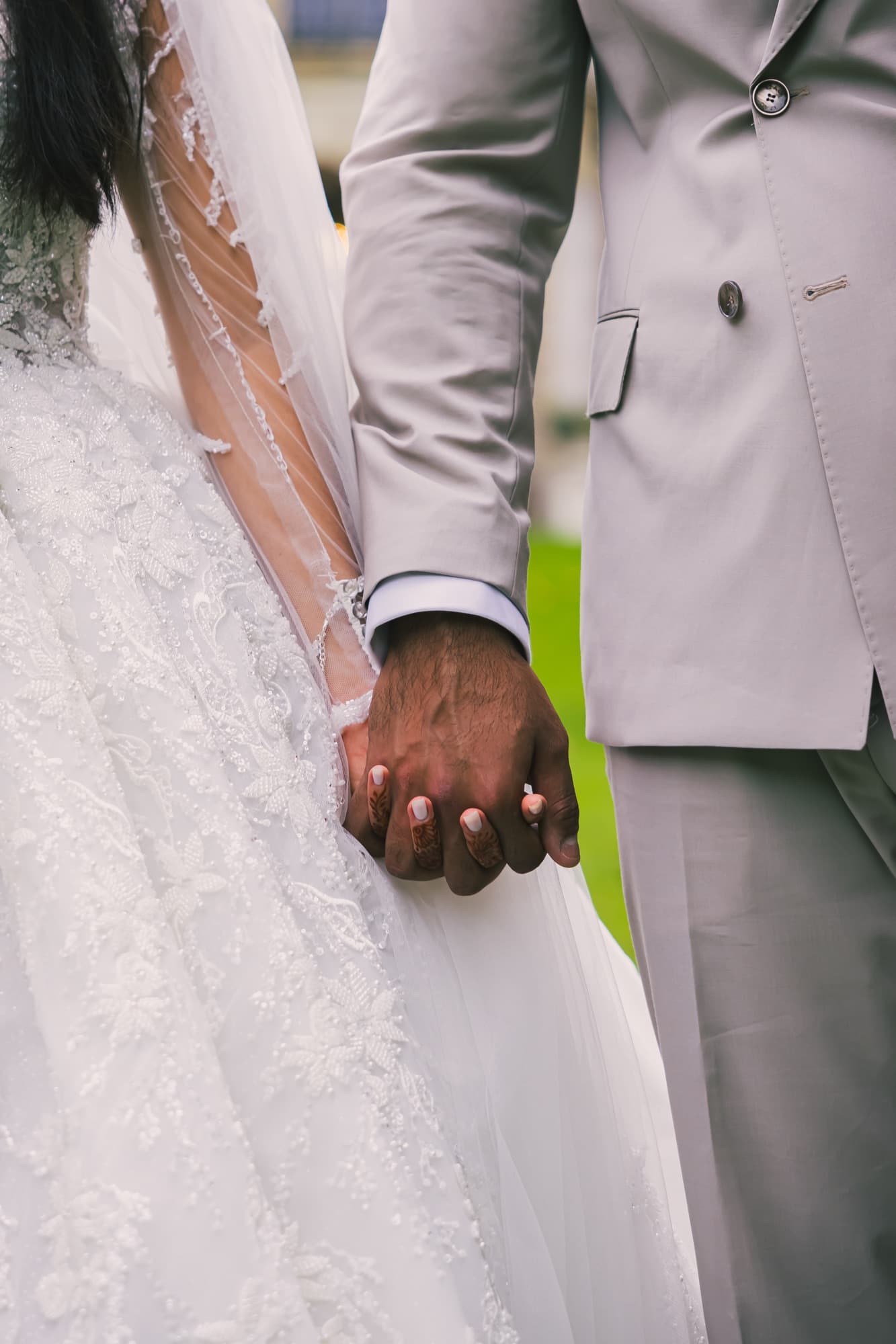 Les mains des mariés enlacées, la robe blanche en dentelle et le costume gris clair, dans l’herbe du parc