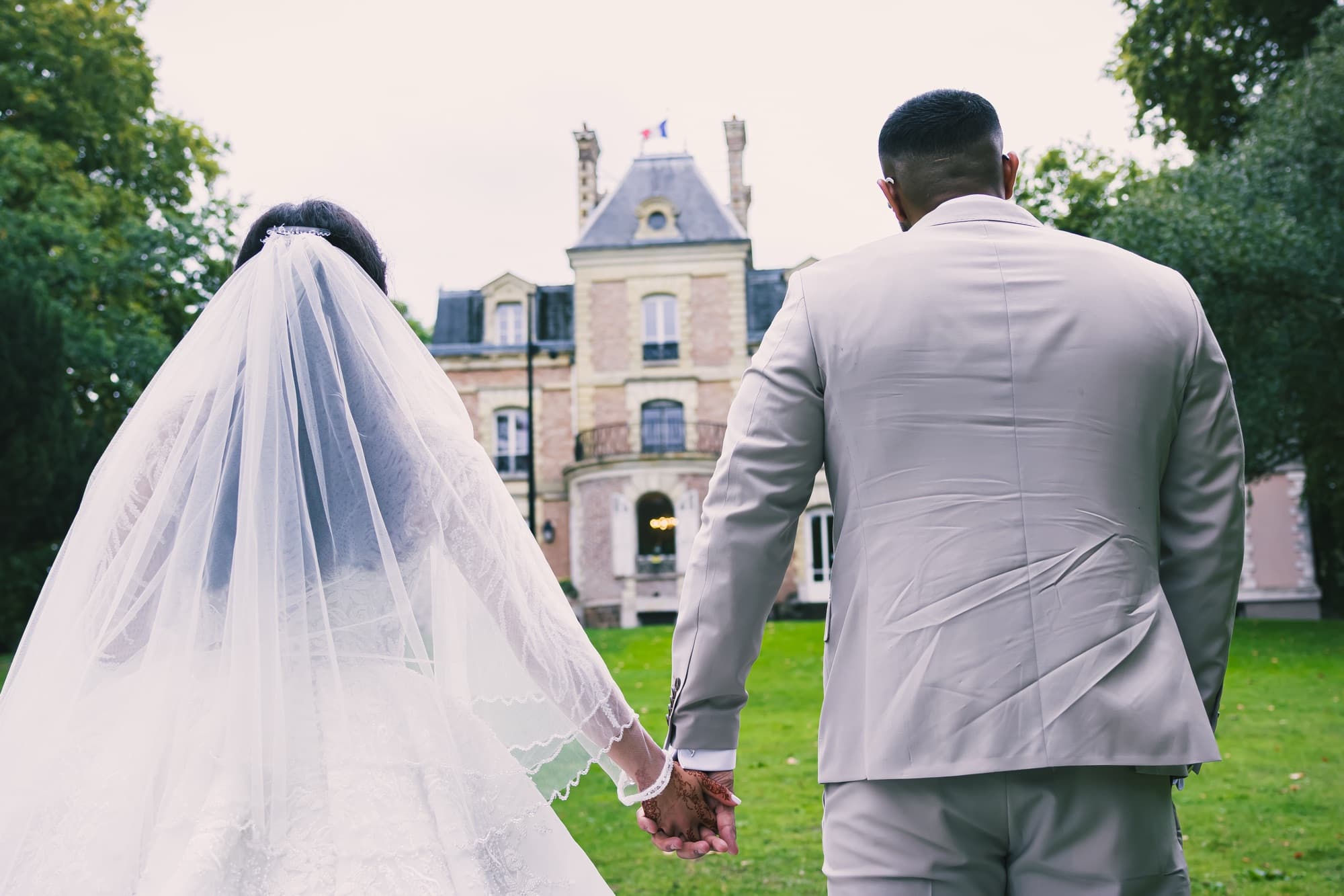 Les mariés de dos dans l’allée du parc, face au château, sous un ciel de pluie fine