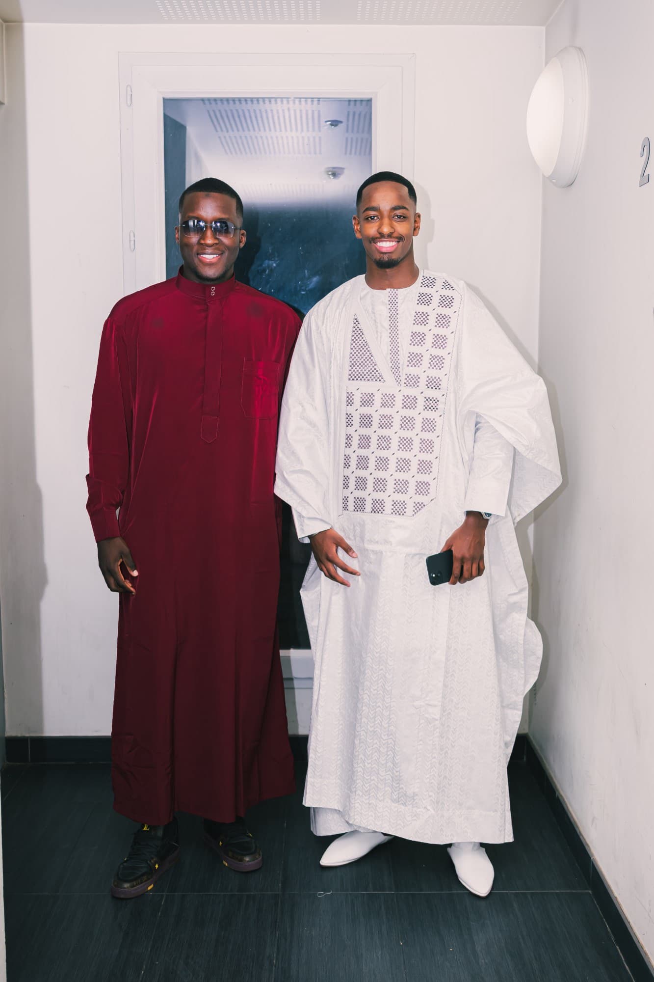 Le marié en tenue blanche brodée posant avec un ami en grand boubou bordeaux, dans un couloir blanc