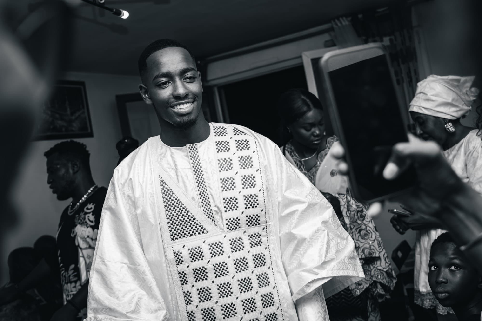 Portrait en noir et blanc du marié en boubou blanc brodé, souriant, au milieu des invités