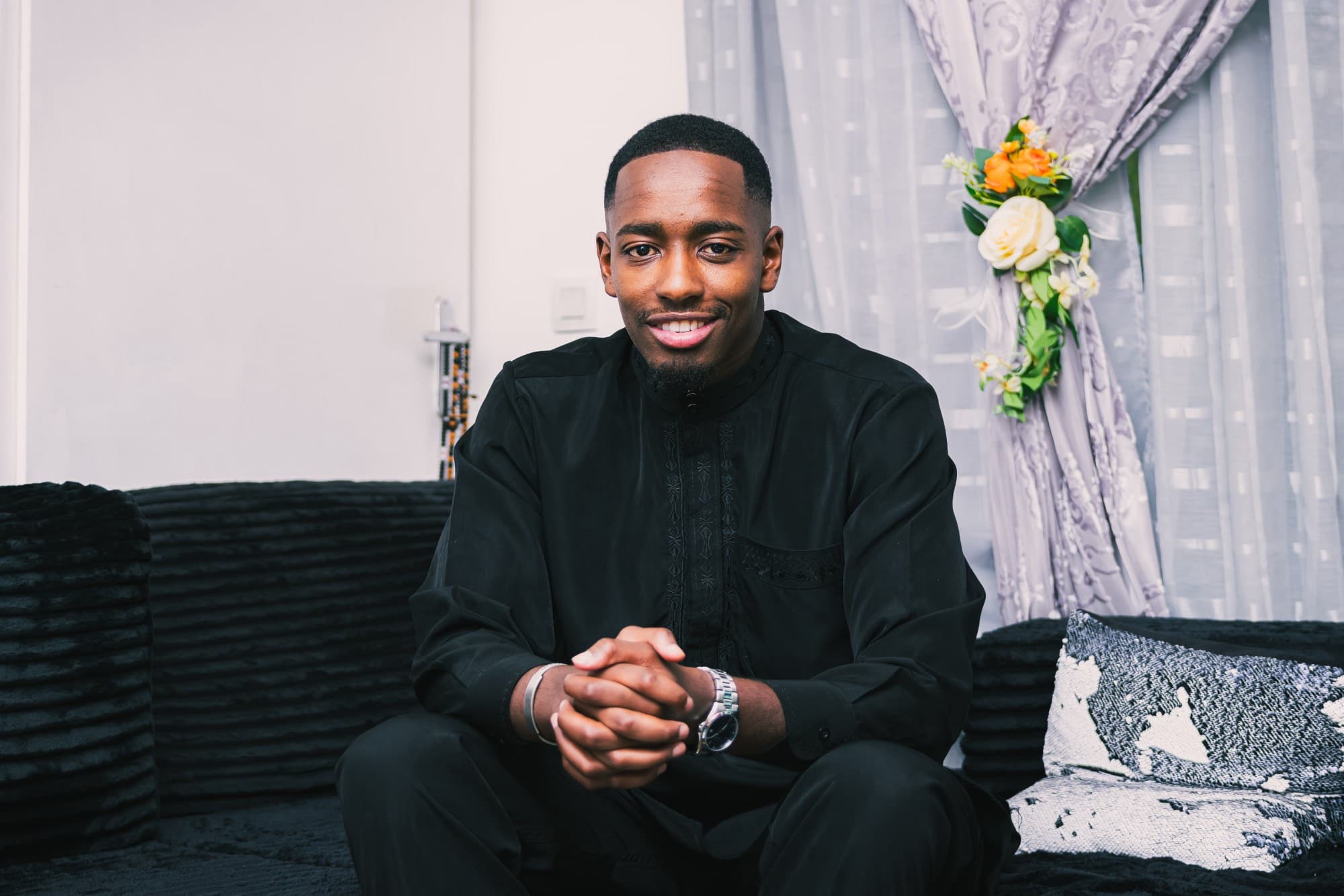 Portrait du marié en tenue noire, assis sur un canapé noir, les mains jointes, devant un rideau fleuri