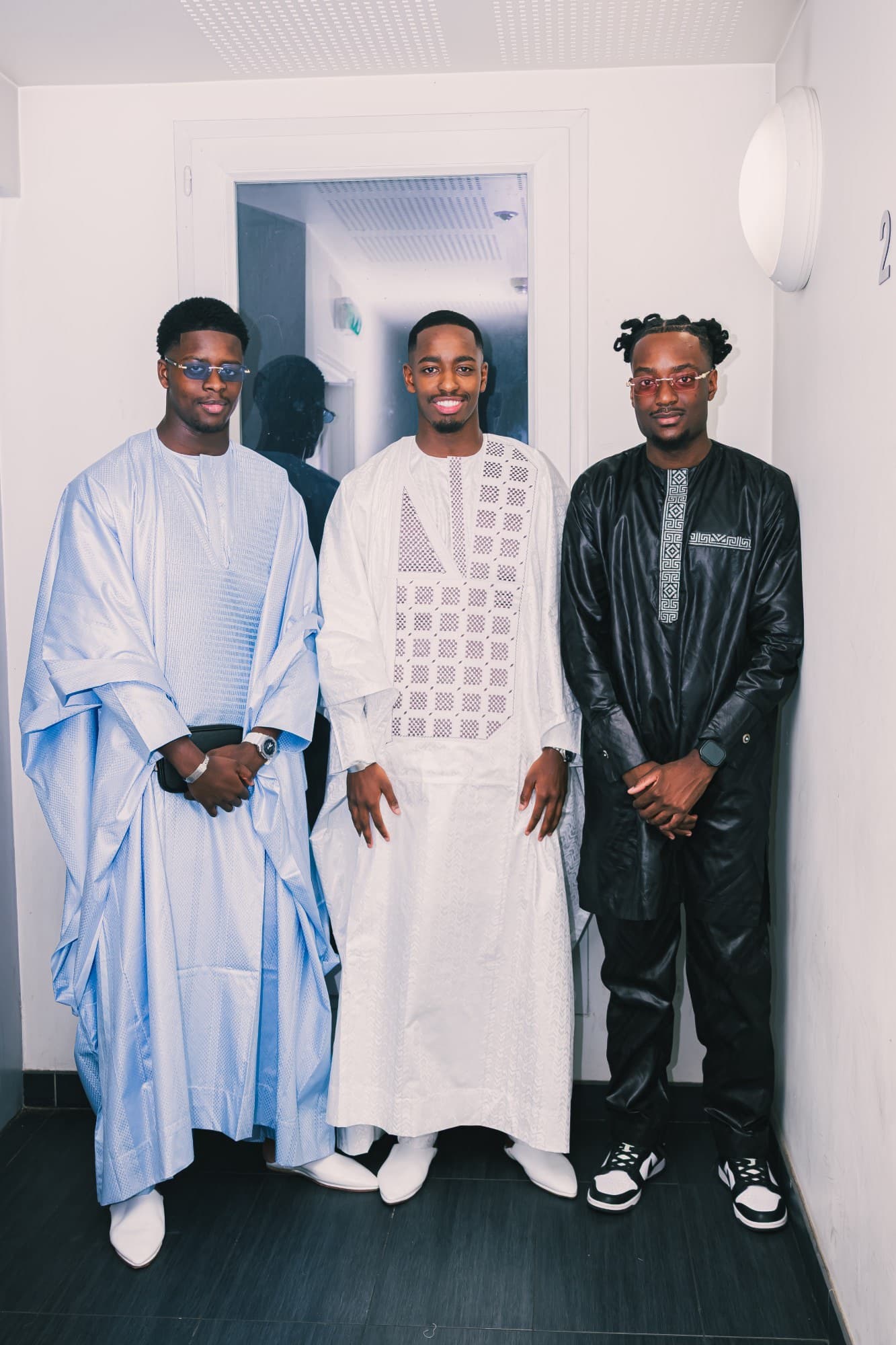 Le marié entouré de deux amis, en boubou bleu clair et en tenue noire brodée, dans un couloir