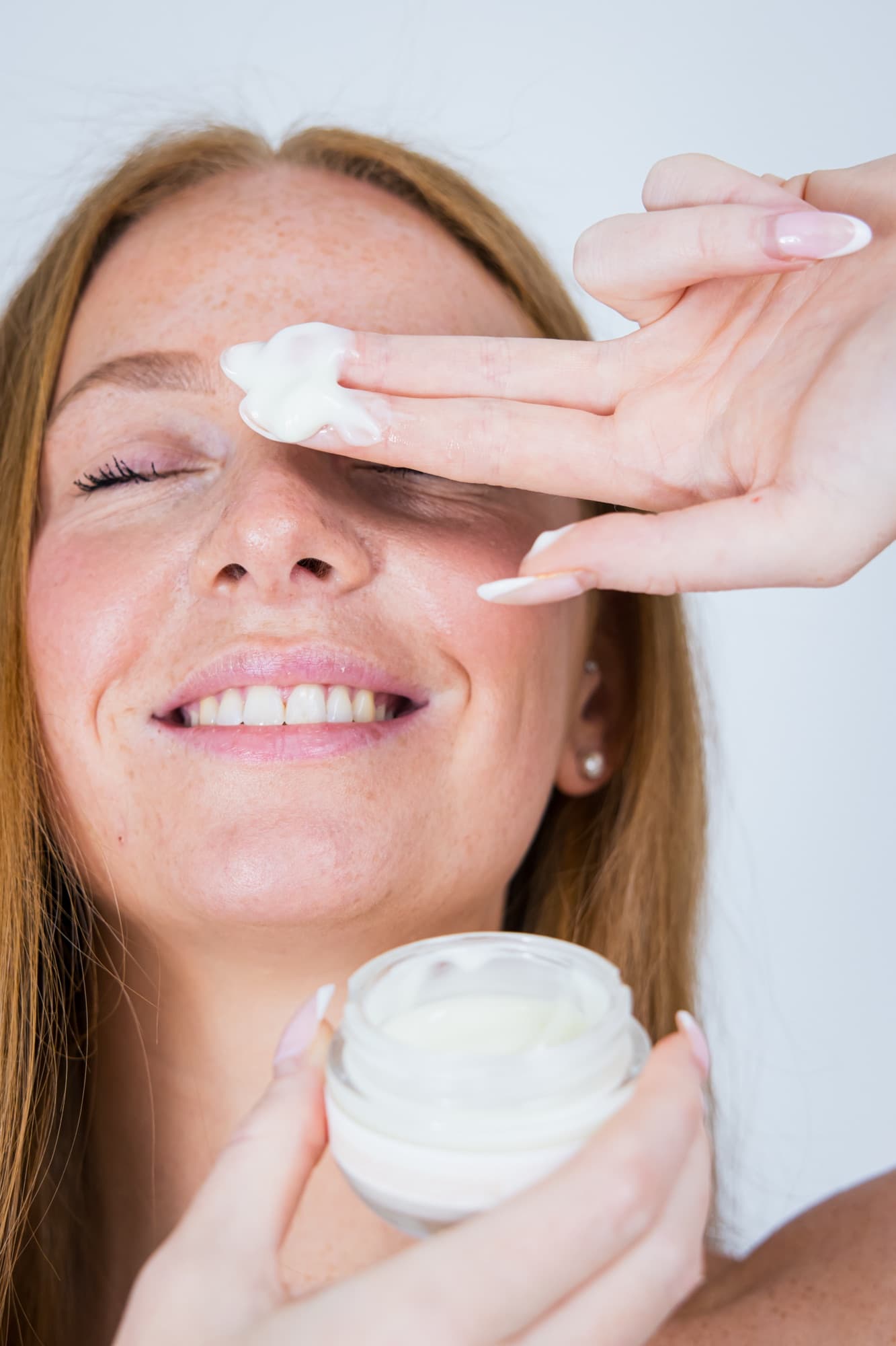 Une jeune femme rousse souriante applique la crème de nuit du bout des doigts, le pot ouvert dans l’autre main