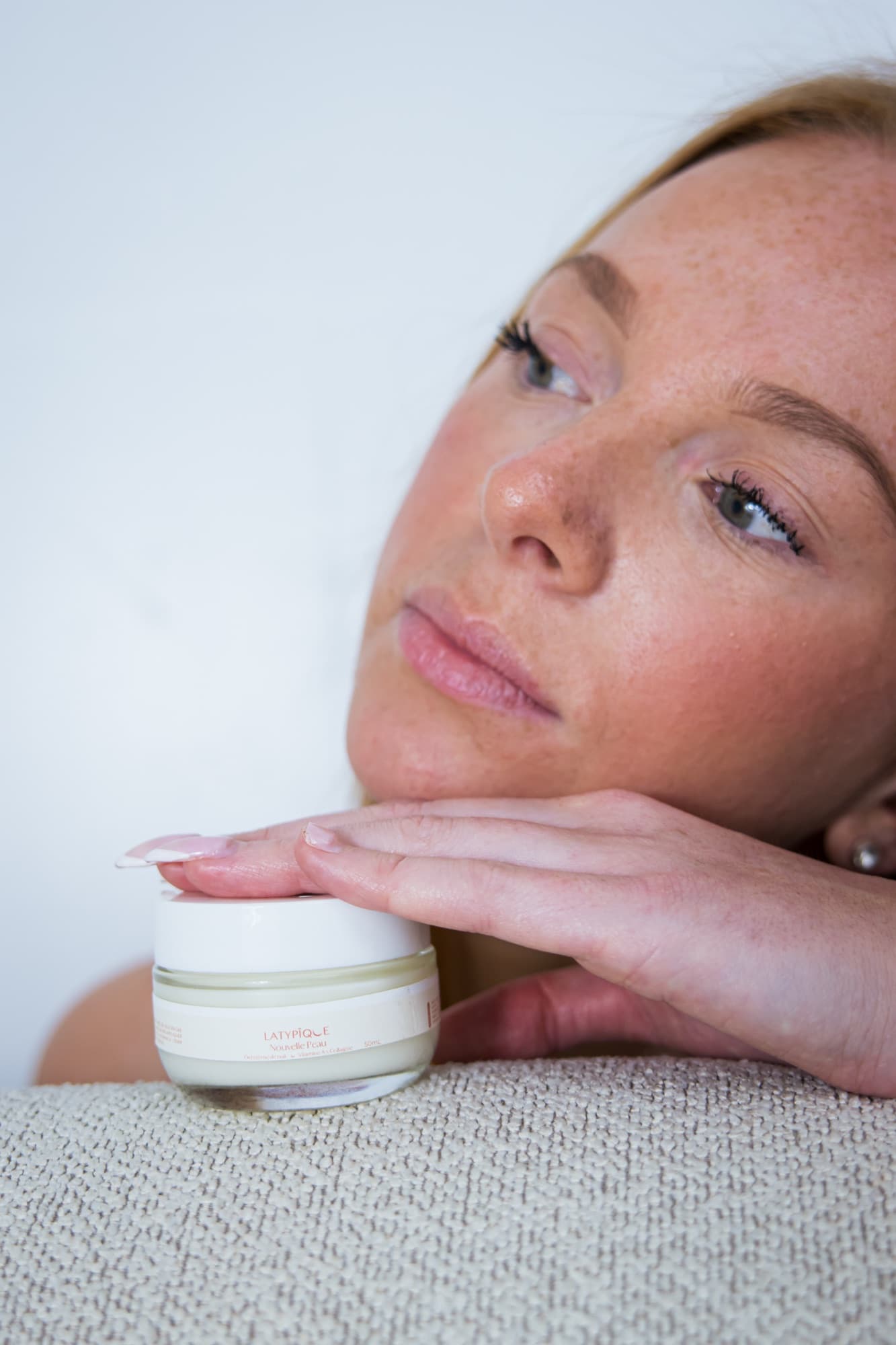 Une femme rousse, la tête appuyée sur ses mains posées sur le pot Nouvelle Peau