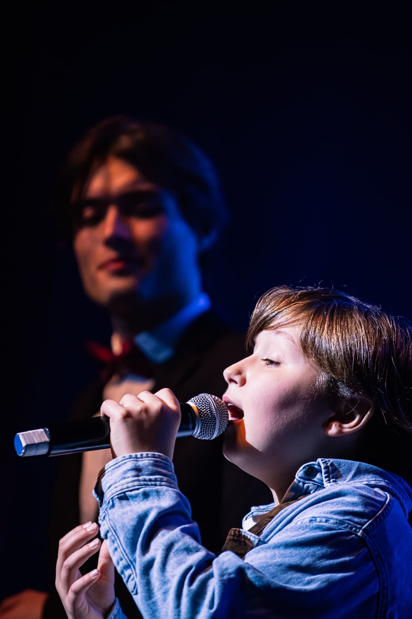 Un jeune garçon chante au micro, un adolescent en nœud papillon derrière lui