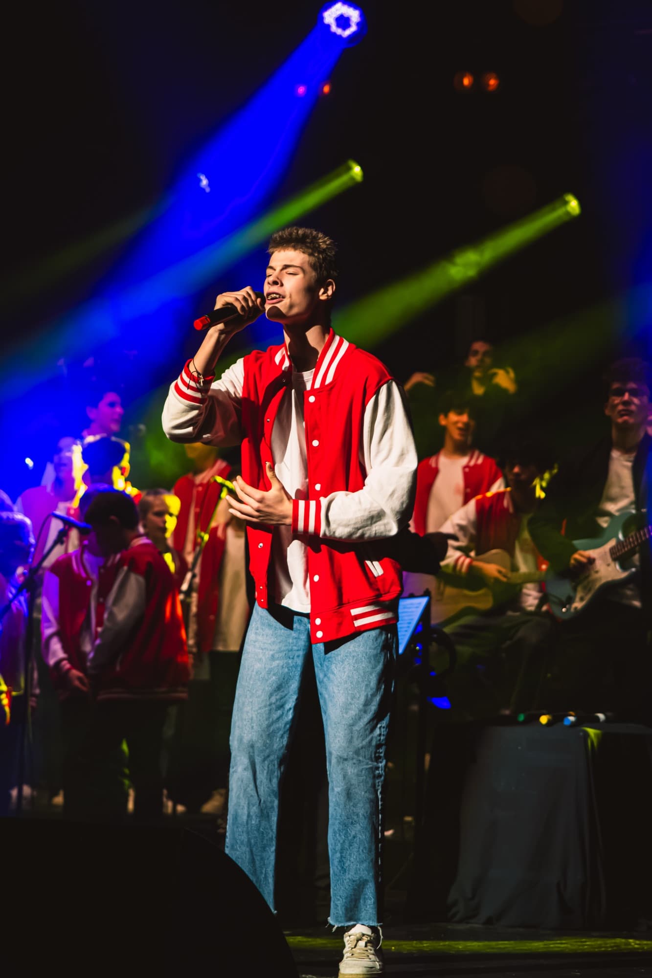 Un chanteur en veste rouge chante devant le groupe, lumières vertes et bleues