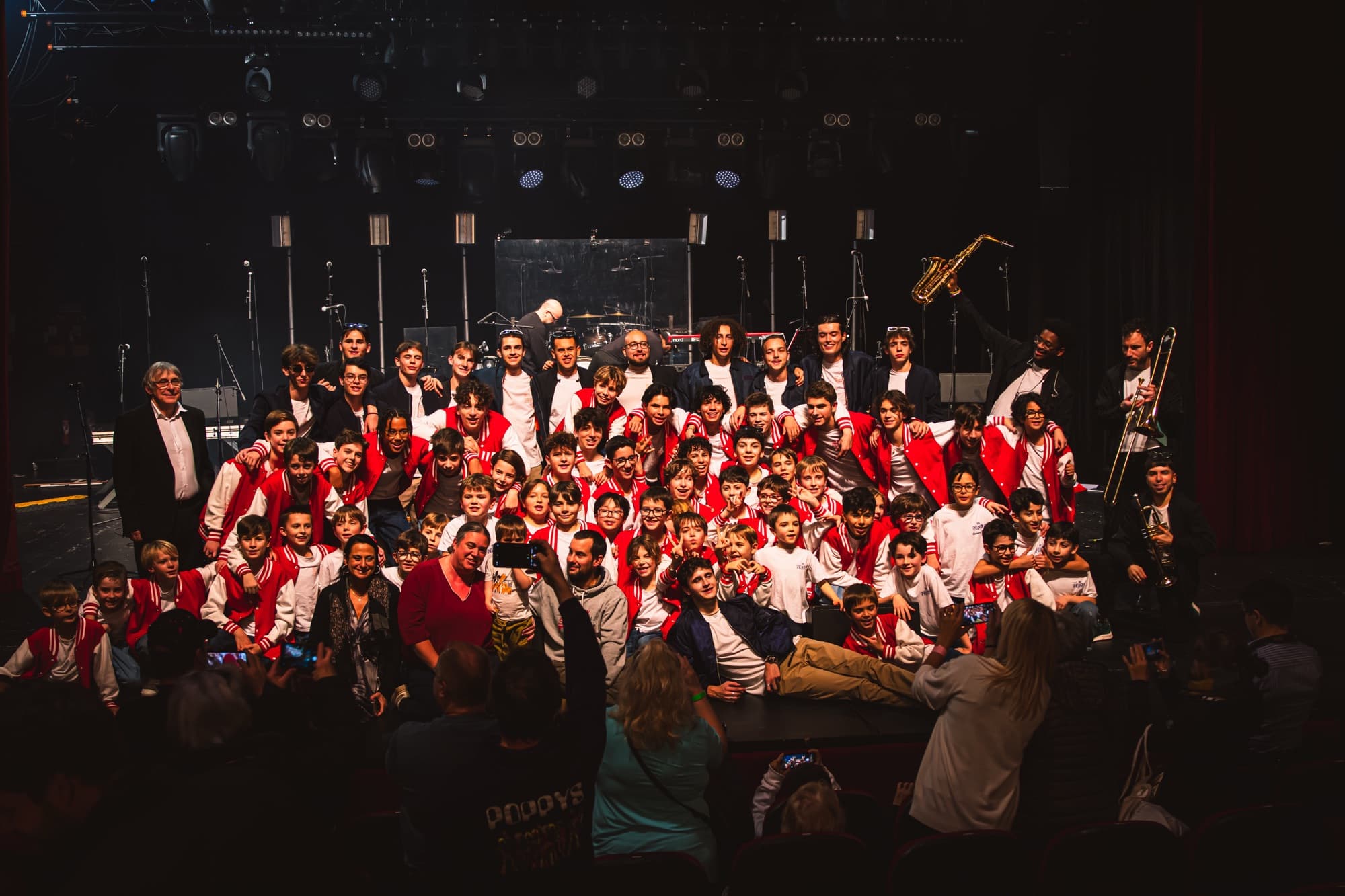 Photo de groupe sur scène à la fin du gala 2025 : les Petits Chanteurs d’Asnières en vestes rouges, les musiciens et l’équipe