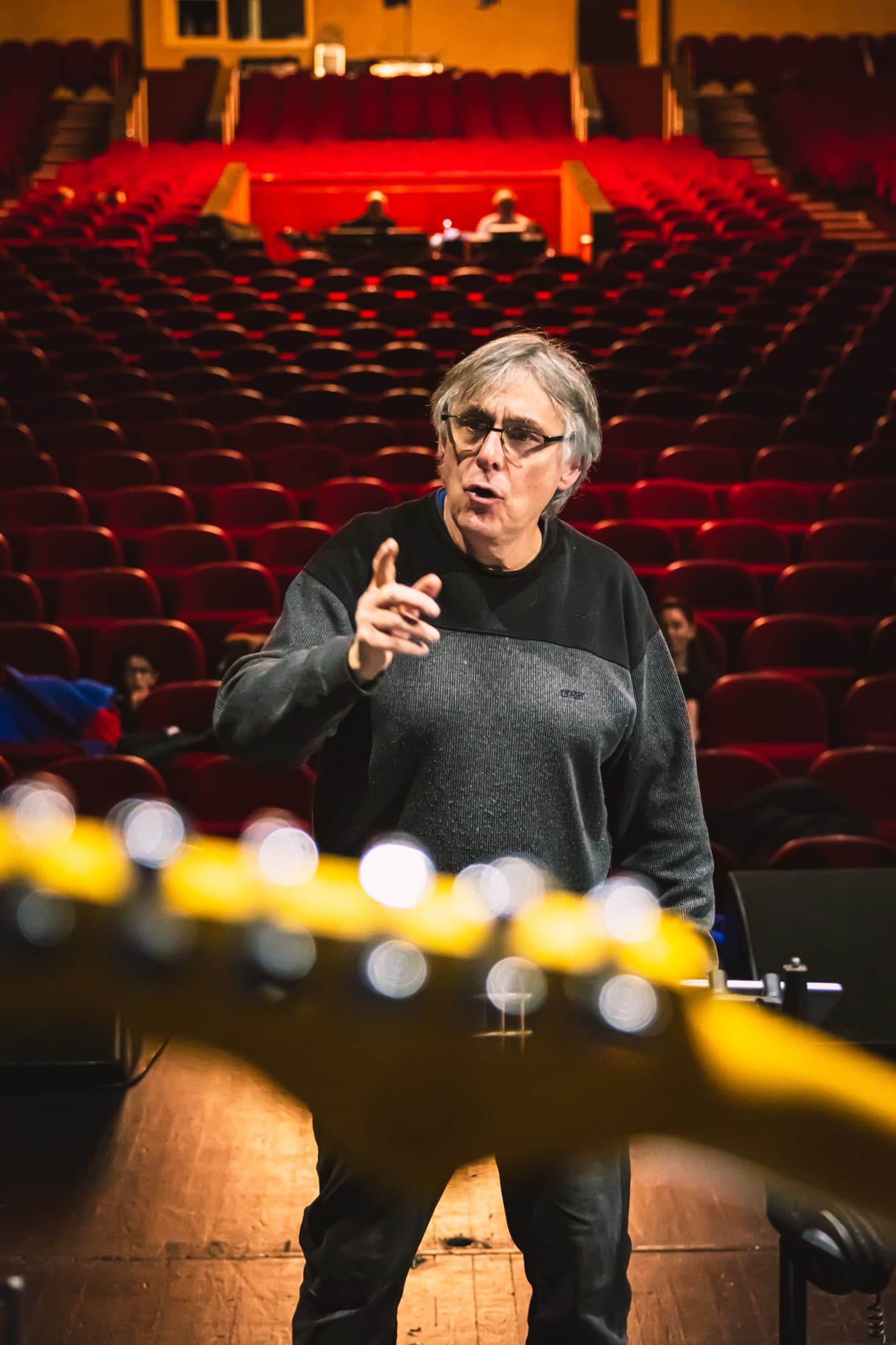 Le chef de chœur en répétition dans la salle vide, un manche de guitare au premier plan
